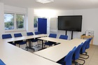 Ein Blick in den Lehrsaal. Man sieht einen hellen Raum mit Tischen, die im Kreis aufgestellt sind. An der Wand stehen ein grßer Monitor, eine Pinnwand und ein Flipchart (Bild anzeigen)